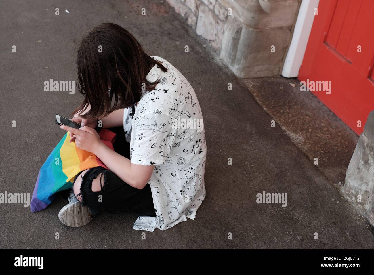 Teen non binario seduto su una piattaforma della stazione ferroviaria e utilizzando il telefono cellulare Foto Stock
