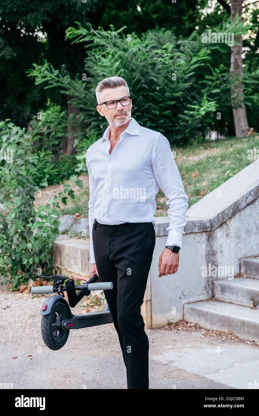 Uomo d'affari senior che tiene in mano uno scooter elettrico mentre si va all'aperto in città Foto Stock