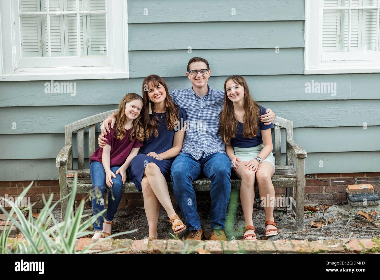 Una famiglia di quattro persone con una madre e un padre e due figlie seduti su una panchina fuori da una casa di cottage blu Foto Stock