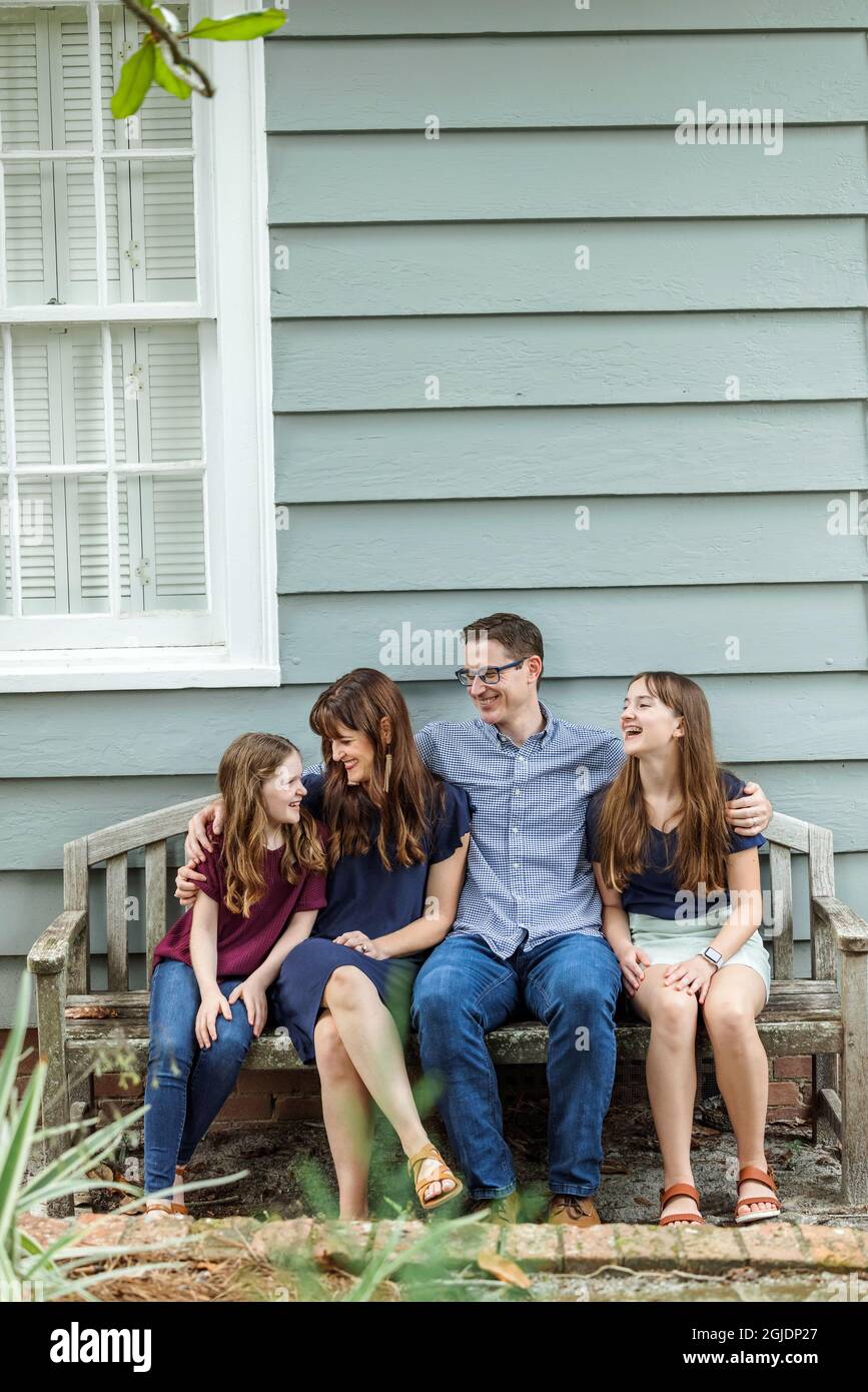 Una famiglia di quattro persone con una madre e un padre e due figlie seduti su una panchina fuori da una casa di cottage blu Foto Stock