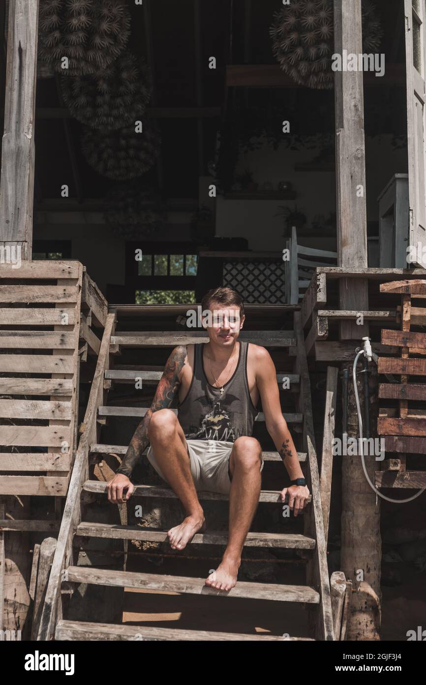 Il giovane uomo caucasico bello con un tatuaggio manica si siede sulle scale di legno di un caffè accogliente era in Asia. Si affaccia sull'oceano. Foto Stock