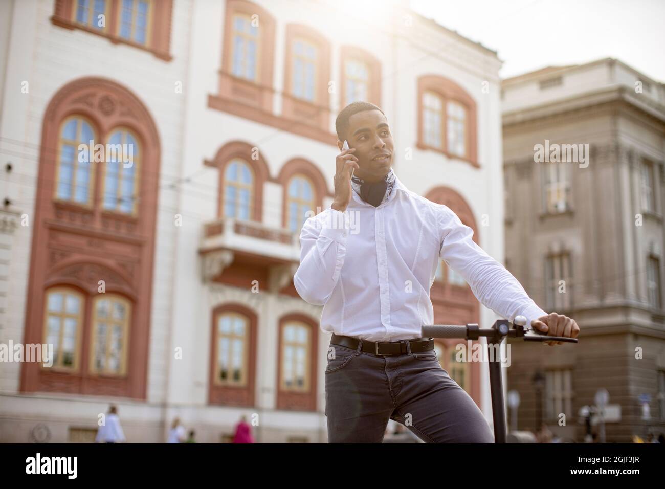 Bel giovane afroamericano che usa il telefono cellulare mentre si è in piedi con uno scooter elettrico su una strada Foto Stock