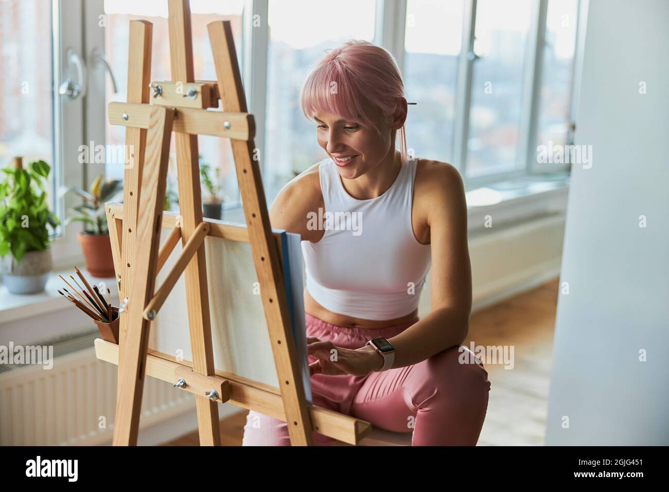 Donna sorridente con capelli rosa si disegna su tela a cavalletto in legno sul balcone Foto Stock
