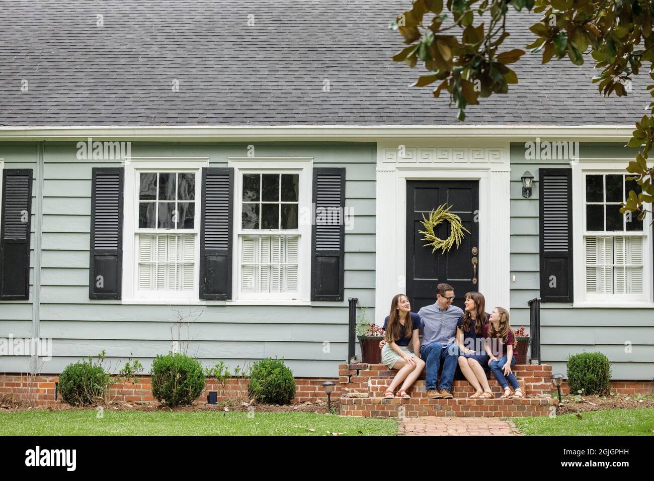 Una famiglia con una madre, un padre e due figlie che si siedono fuori sui gradini di mattone di un portico anteriore di una piccola casa di cottage blu Foto Stock