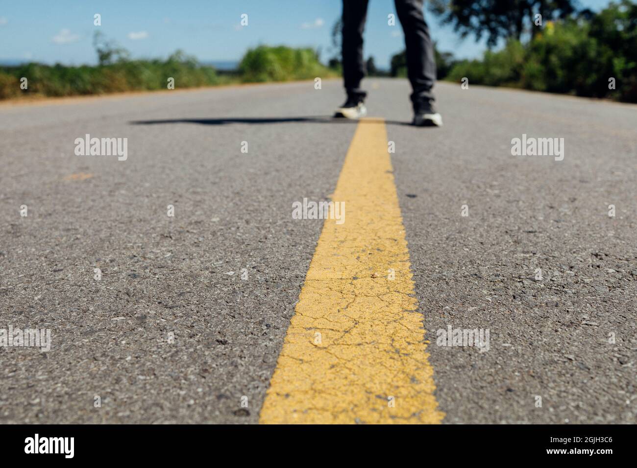 Piedi da uomo in piedi su strada asfaltata con linee di marcatura gialle. Uomo che indossa scarpe da ginnastica e jeans. Foto Stock