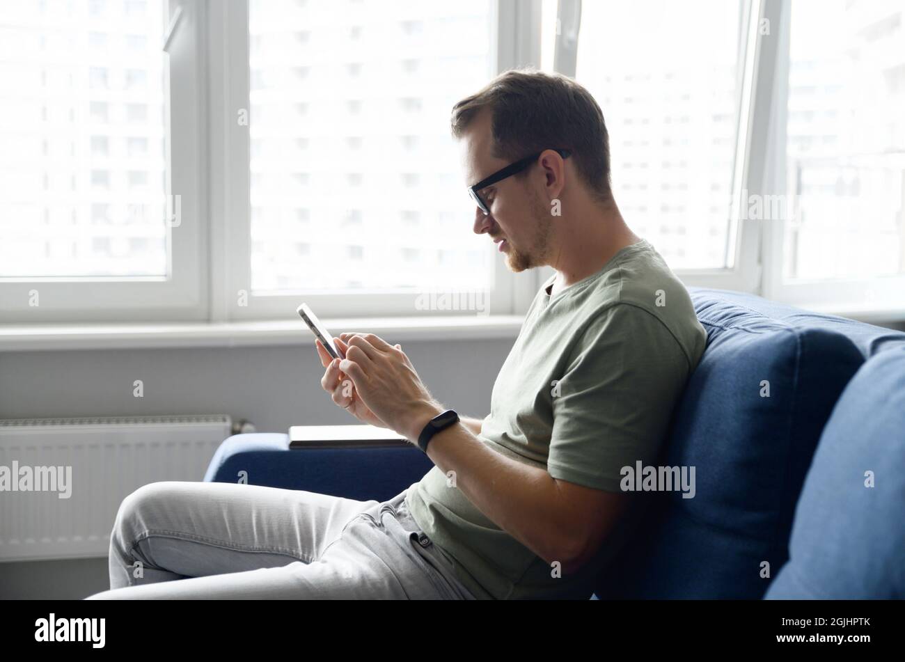 Ragazzo sorridente che indossa un abbigliamento casual, con messaggi SMS sullo smartphone. Vista laterale un giovane si siede sul divano moderno e utilizza l'app mobile, le notizie dei feed di scorrimento, il websurf, il gioco Foto Stock