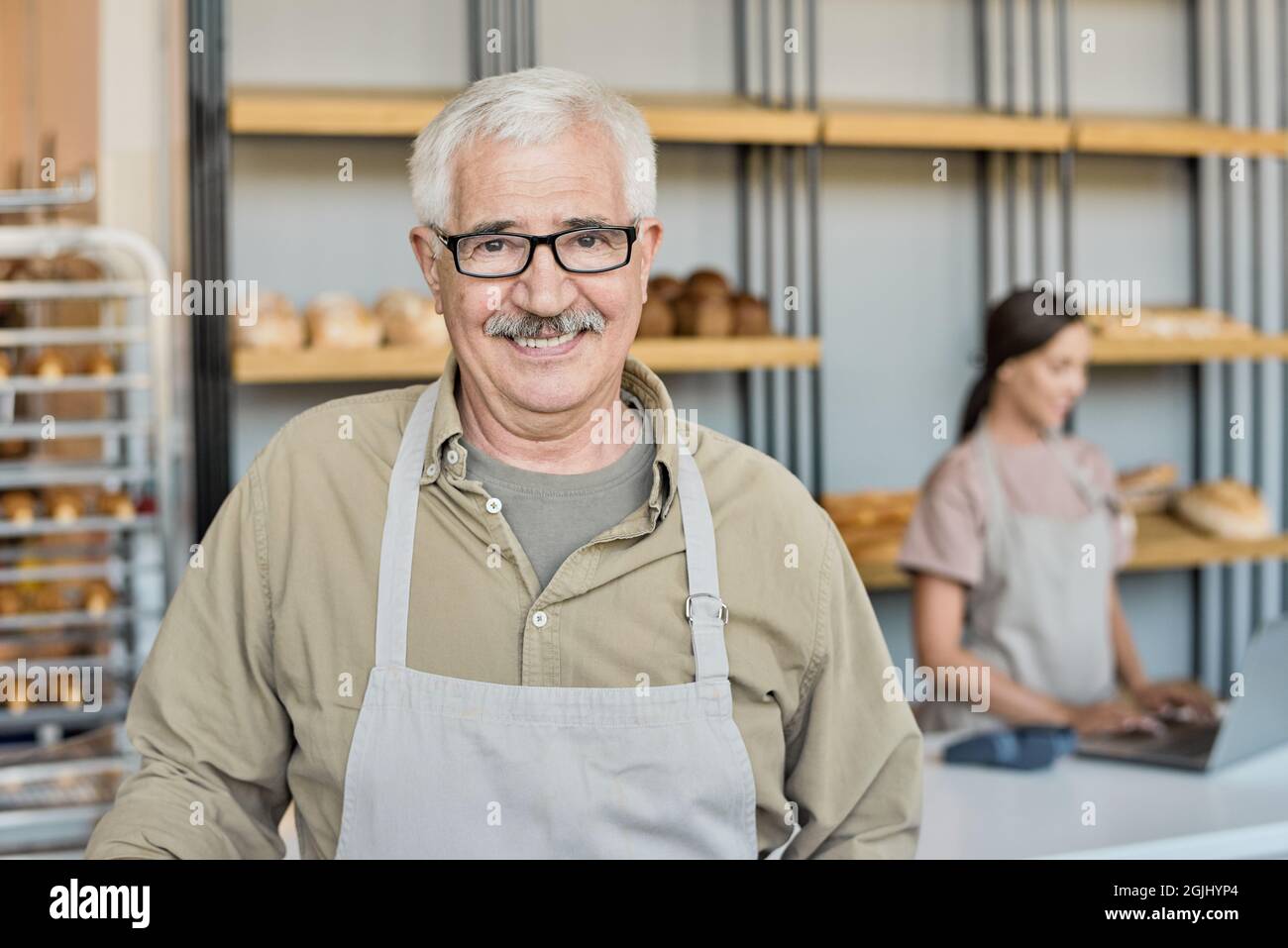 Ritratto di uomo maturo sorridente in grembiule e occhiali in piedi contro esposizione con pane fresco in panetteria casa Foto Stock