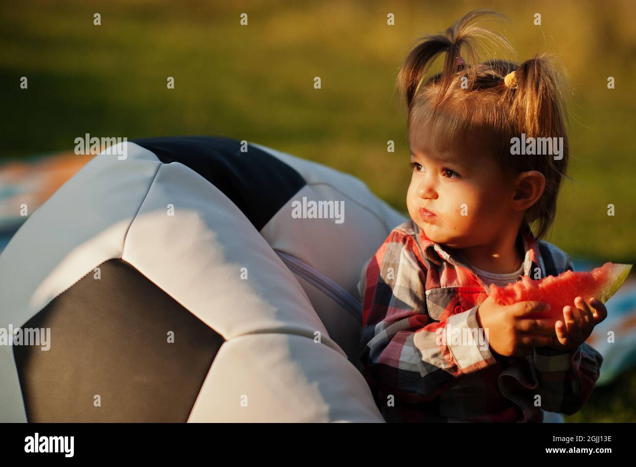 Carina bambina in camicia a scacchi sedersi sul pallone da calcio pouf e mangiare l'anguria. Foto Stock