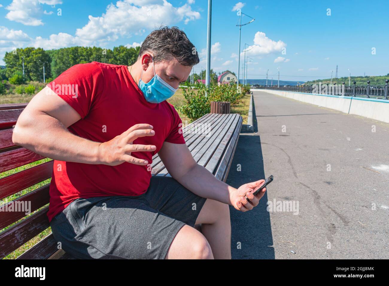 L'uomo infastidito che indossa una maschera medica comunica emotivamente tramite comunicazione video mentre si siede su una panca del parco Foto Stock