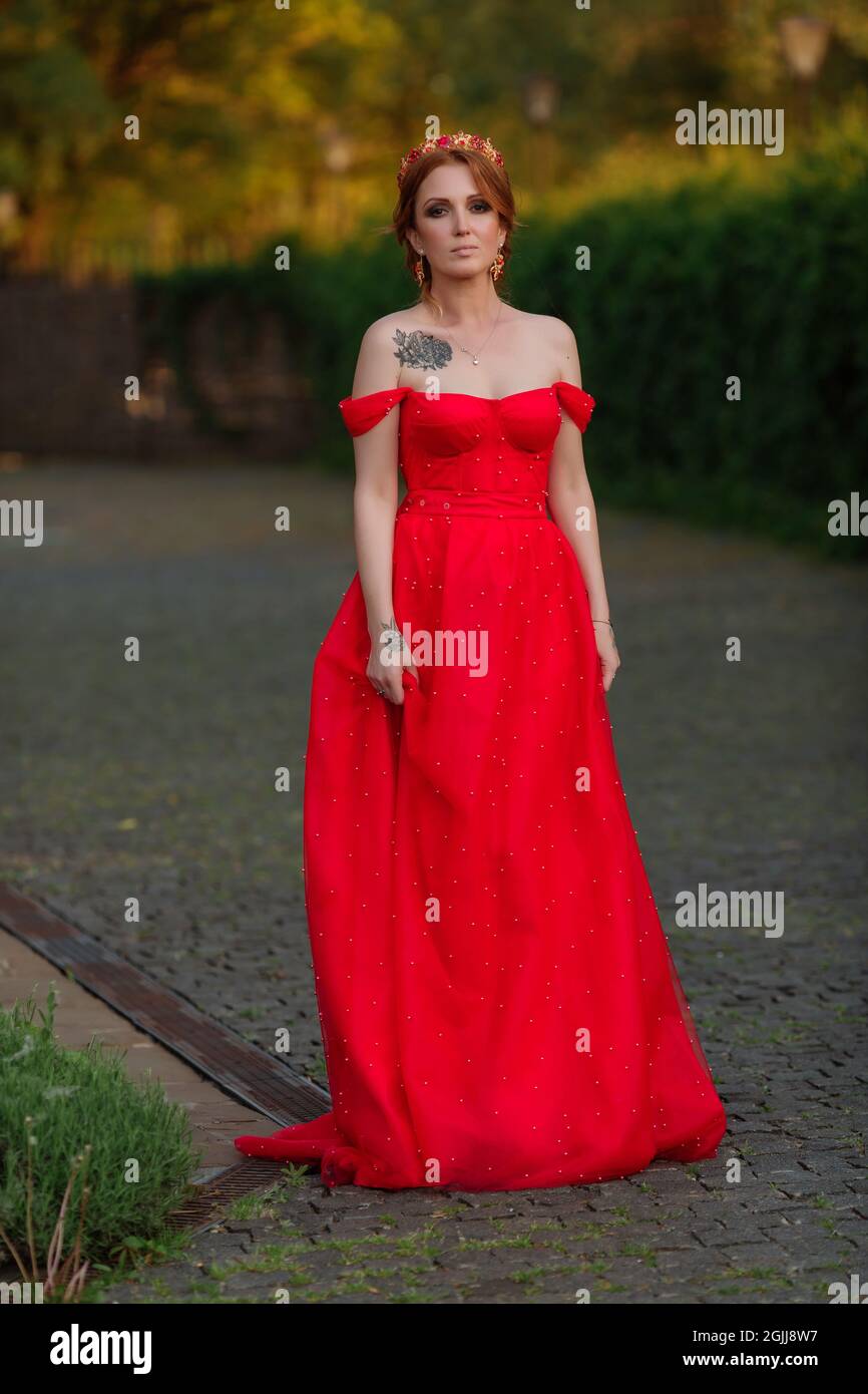 Bella donna tatuata rossa in lungo vestito rosso e diadema sulla testa  camminando sullo sfondo del castello medievale offuscato al tramonto estivo  Foto stock - Alamy