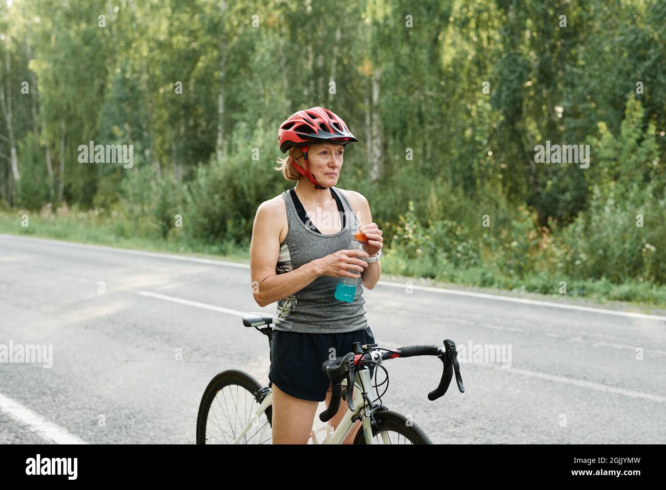 Donna in casco in piedi sulla strada e bere acqua dopo il suo allenamento in bicicletta Foto Stock