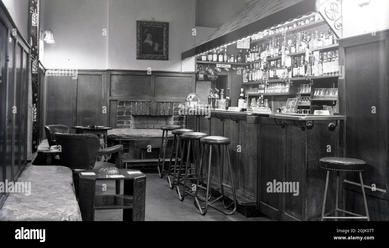 Anni '50, storico, all'interno di un pub bar tradizionale, Witney, Oxford, Inghilterra, Regno Unito. Conosciuto anche come un bar accogliente, ha un'atmosfera intima con estesi pannelli in legno, sgabelli in metallo e altri posti a sedere. Posacenere sul bancone bar e tavoli bassi Foto Stock
