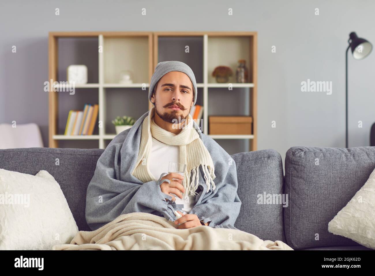 Giovane malato in abiti caldi seduto con un bicchiere d'acqua e spiegare i sintomi al medico online Foto Stock