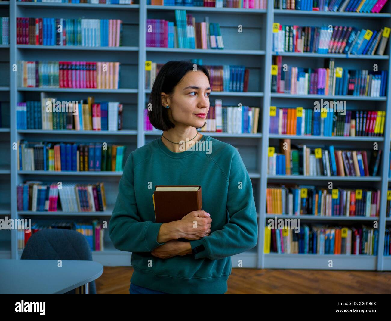 Giovane e attraente studentessa donna legge un libro che si prepara per un esame mentre sullo sfondo di scaffali. Foto Stock