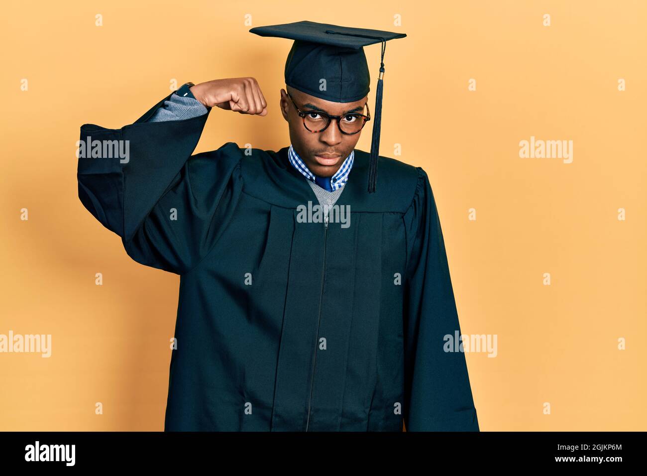 Il giovane afroamericano che indossa il cappuccio di graduazione e la cerimonia vestisce la persona forte che mostra il muscolo del braccio, fiducioso e fiero di potere Foto Stock