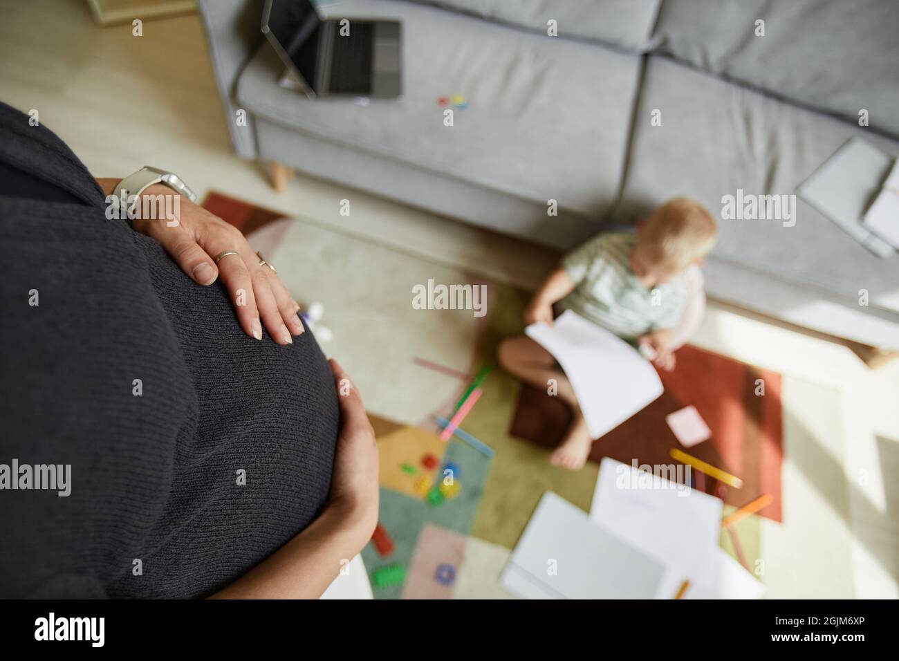 Sopra la vista di donna incinta irriconoscibile che struca il ventre mentre suo figlio è seduto su tappeto e disegno Foto Stock