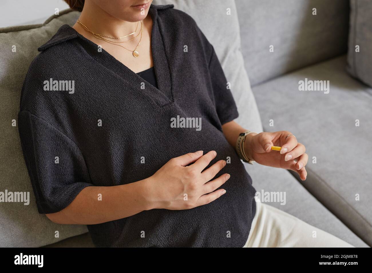 Primo piano di una donna incinta in camicia nera seduta sul divano e prendere la vitamina mentre si strofinano il ventre Foto Stock