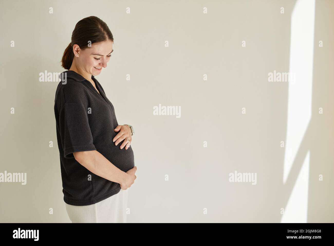 Felice bella incinta giovane donna caucasica con ponytail che è in previsione del bambino abbracciare il ventre Foto Stock