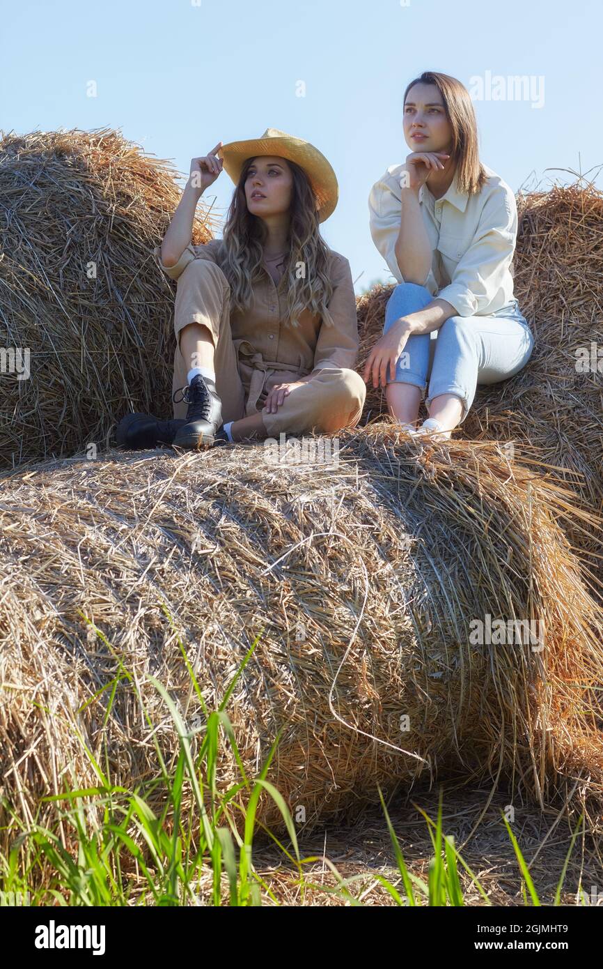 Due giovani donne bruna e bionda sono sedute sui rotoli di fieno. Romantiche ragazze di campagna sul fieno. Foto Stock