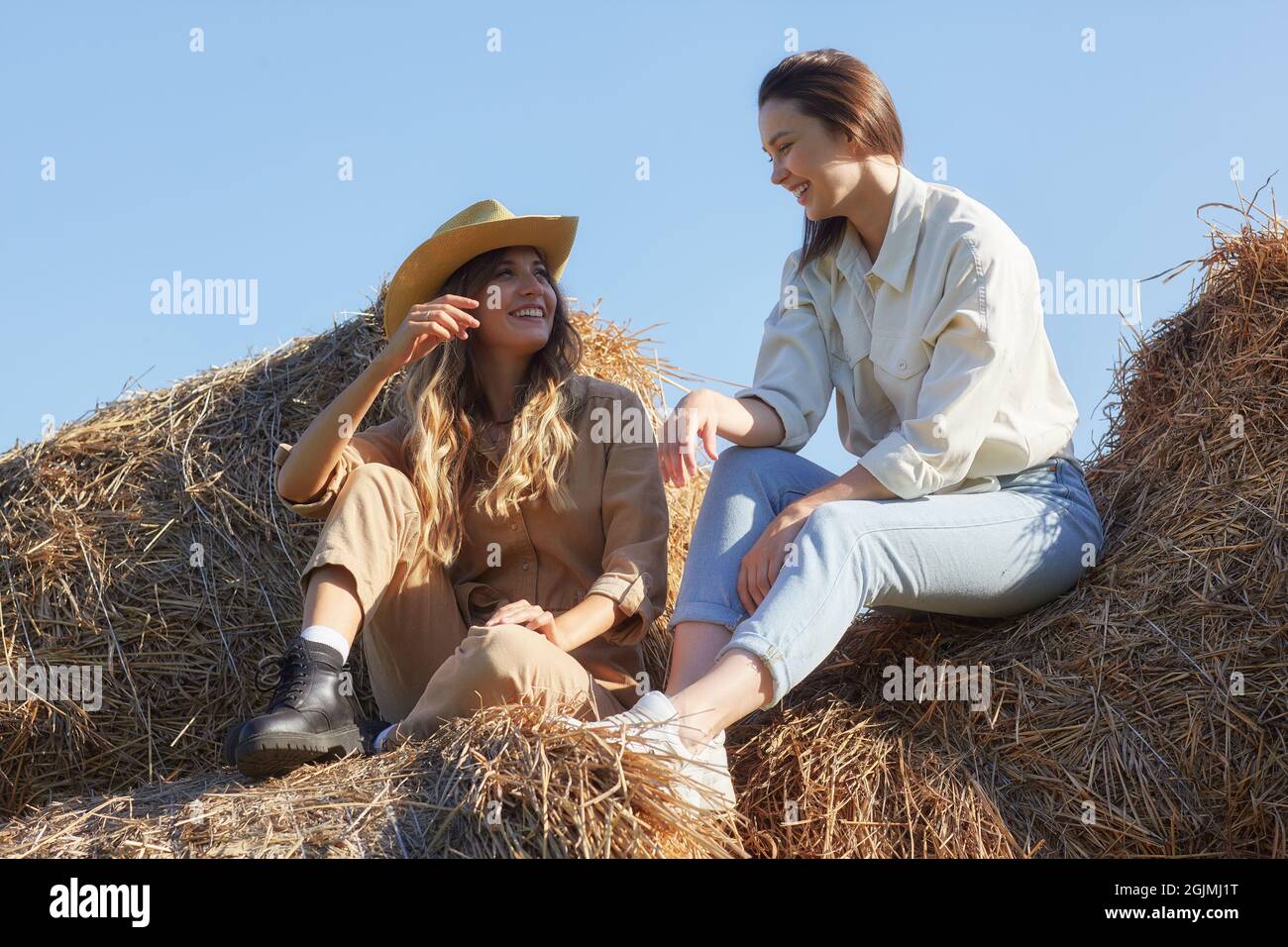Due giovani donne bruna e bionda sono sedute sui rotoli di fieno. Romantiche ragazze di campagna sul fieno stanno parlando e ridendo. Foto Stock