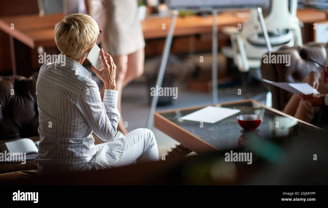 Una donna d'affari anziana seduta sul divano e che parla sullo smartphone mentre ascolta una presentazione di una collega femminile in un'atmosfera piacevole Foto Stock