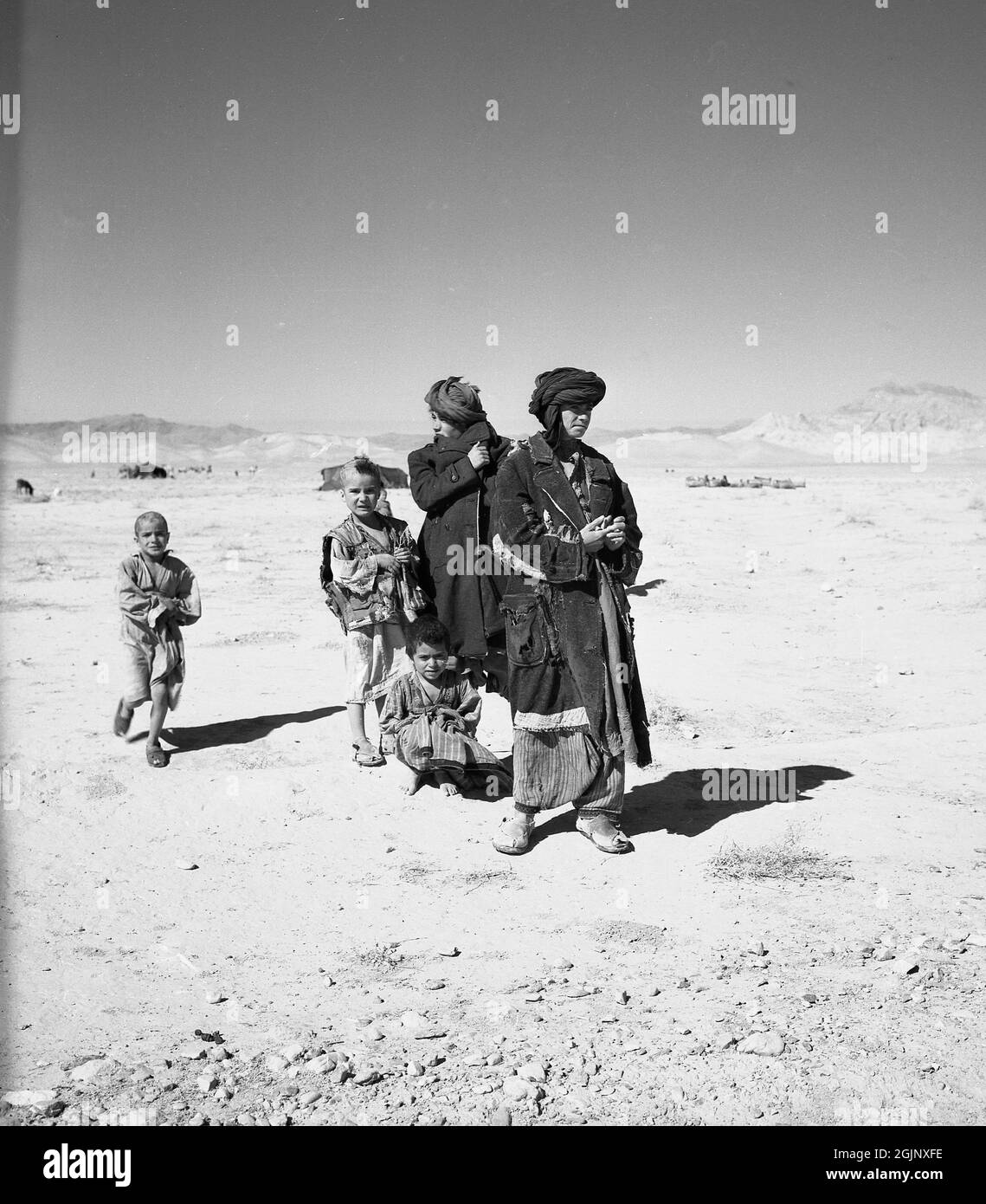 Anni '50, storico, due giovani uomini afghani nomadi che indossano torbani e vestiti stracciati in piedi con bambini piccoli fuori nel deserto arido e arido dell'Afghanistan. Foto Stock