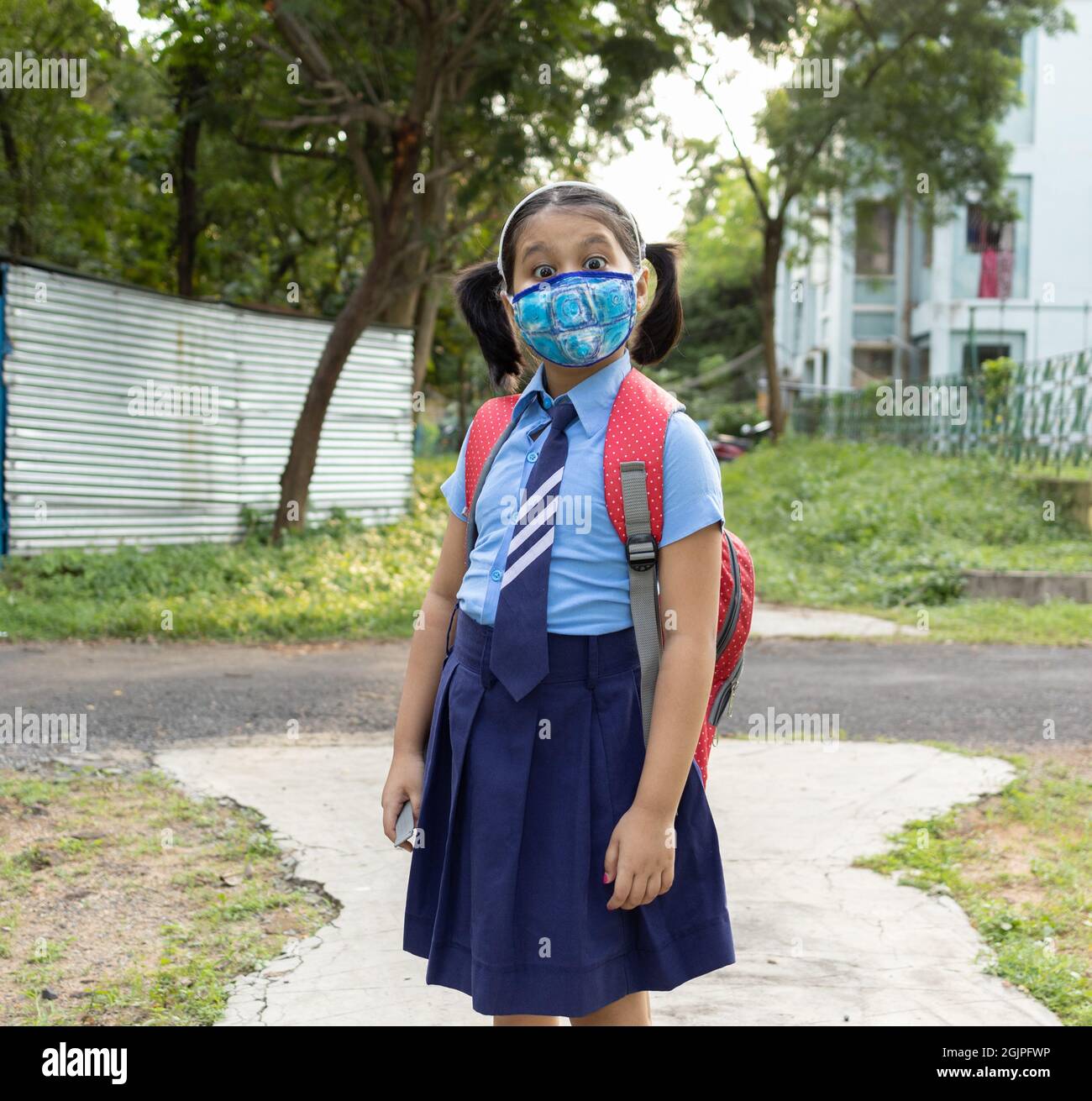 Ritratto di una ragazza indiana felice studente bambino in uniforme blu scuola con sacco rosso e maschera naso protezione andare a scuola Foto Stock
