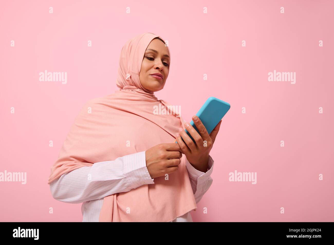 Donna musulmana matura di etnia mediorientale in hijab rosa con smartphone in copertina blu sulle mani, isolata su sfondo rosa pastello con copia Foto Stock