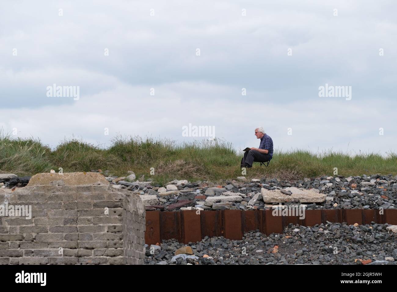 Un artista seduto sul sito dell'ex centrale elettrica di Carmarthen Bay, che si affaccia su una spiaggia al Porto di Burry. I resti dell'edificio industriale demolito, la spiaggia e l'amianto sono stati scoperti dall'erosione delle maree. I segnali di avvertimento sono stati posti nelle vicinanze dall'autorità locale. Foto Stock