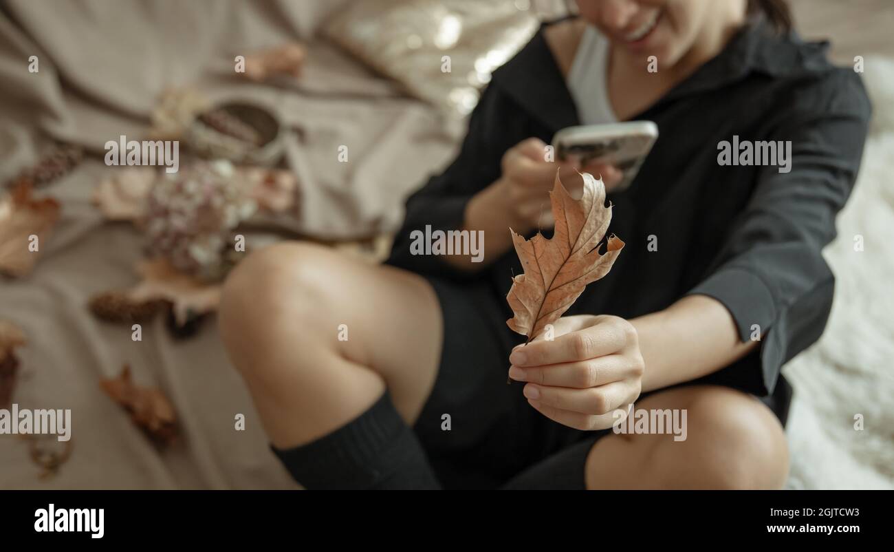 Una giovane donna in calze a maglia utilizza il telefono in un letto accogliente, tra le foglie autunnali. Foto Stock