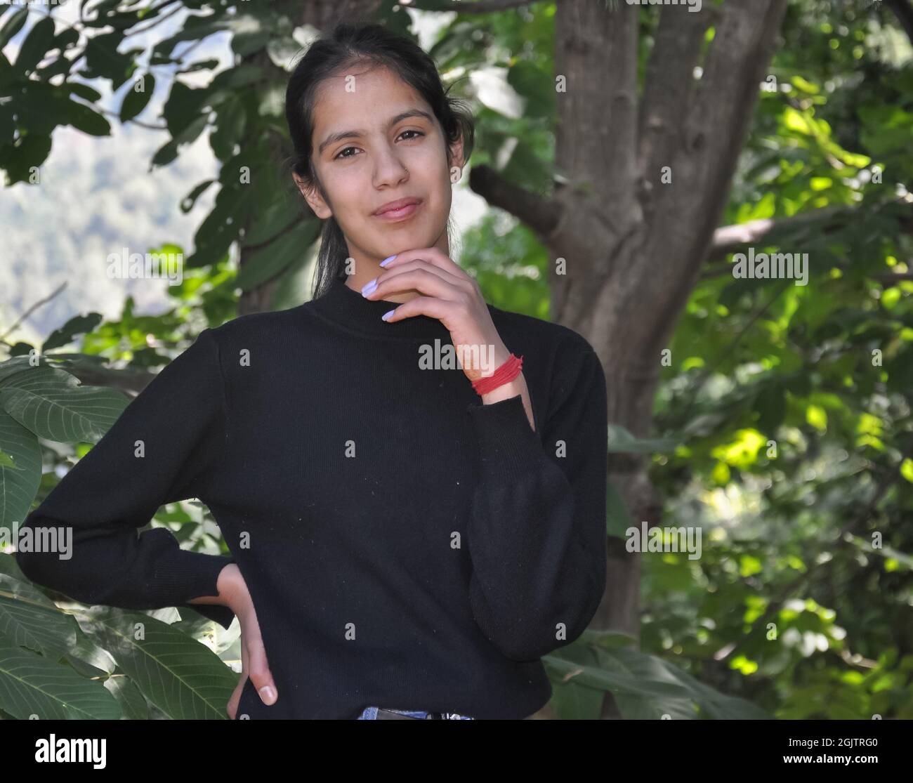 Primo piano ritratto di una bella nord indiana giovane ragazza in piedi in natura posando con una mano sul mento e l'altra sui fianchi con guardare la macchina fotografica Foto Stock