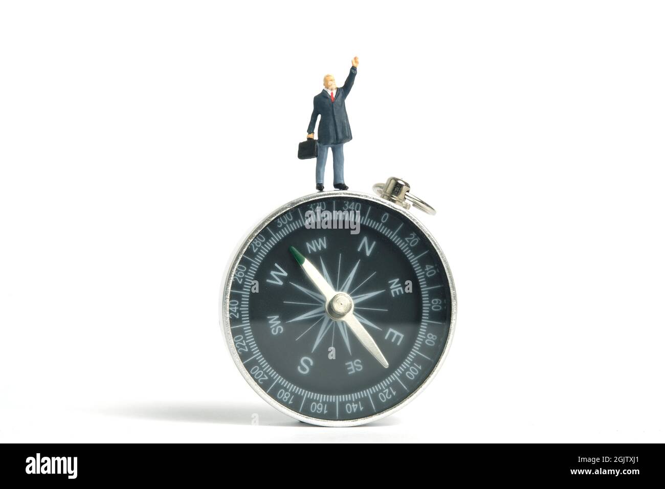 Fotografia di personaggi giocattolo in miniatura. Concetto di direzione aziendale. Un uomo d'affari in piedi sopra una bussola nera mentre alza la mano. Isolato su wh Foto Stock