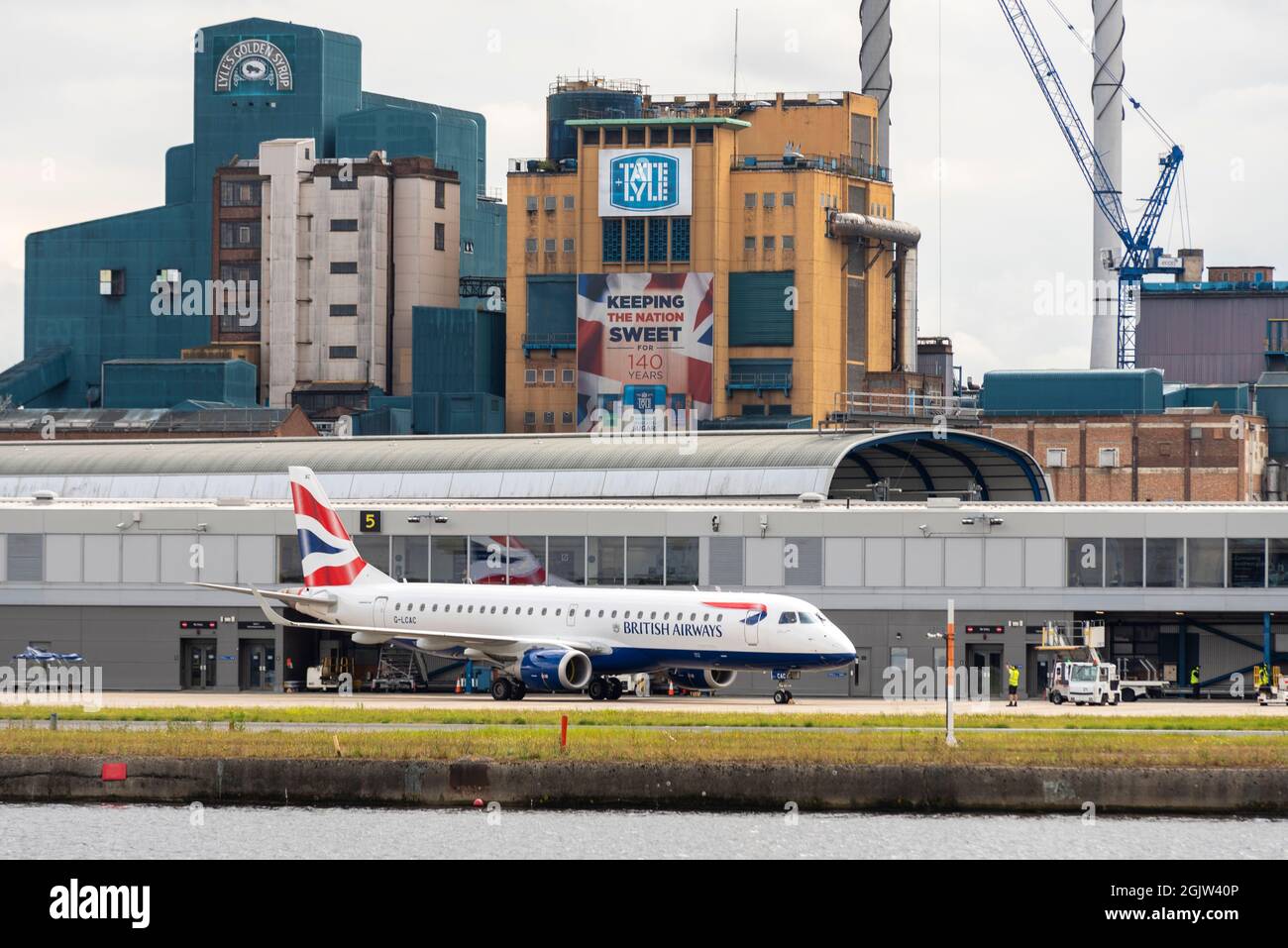 London City Airport, con l'iconica fabbrica di raffinerie di zucchero Tate & Lyle alle spalle. British Airways Cityflyer Embraer ERJ 190 aereo jet in stand Foto Stock