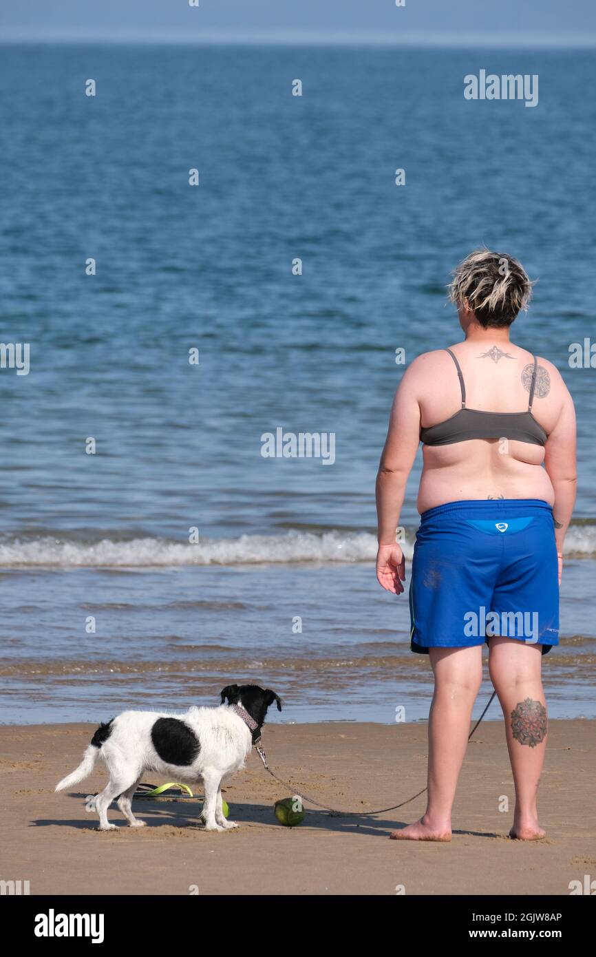 Una giovane donna tatuata con un piccolo cane in piedi sulla riva di Broughton Beach, penisola di Gower, Galles, Regno Unito. Foto Stock