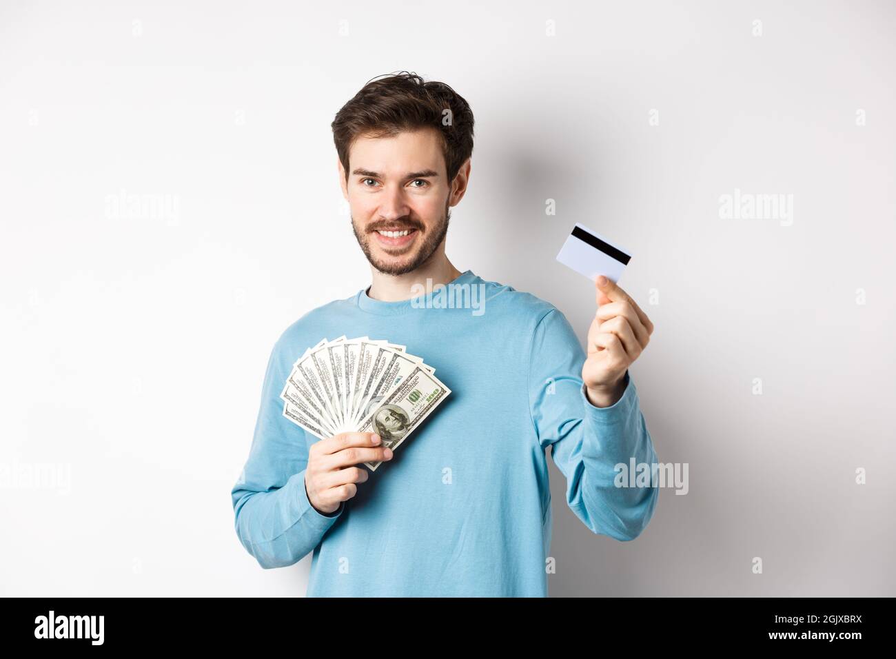 Bel giovane uomo sorridente e che offre il pagamento in contanti e senza contatto, mostrando denaro con carta di credito in plastica, in piedi su sfondo bianco Foto Stock