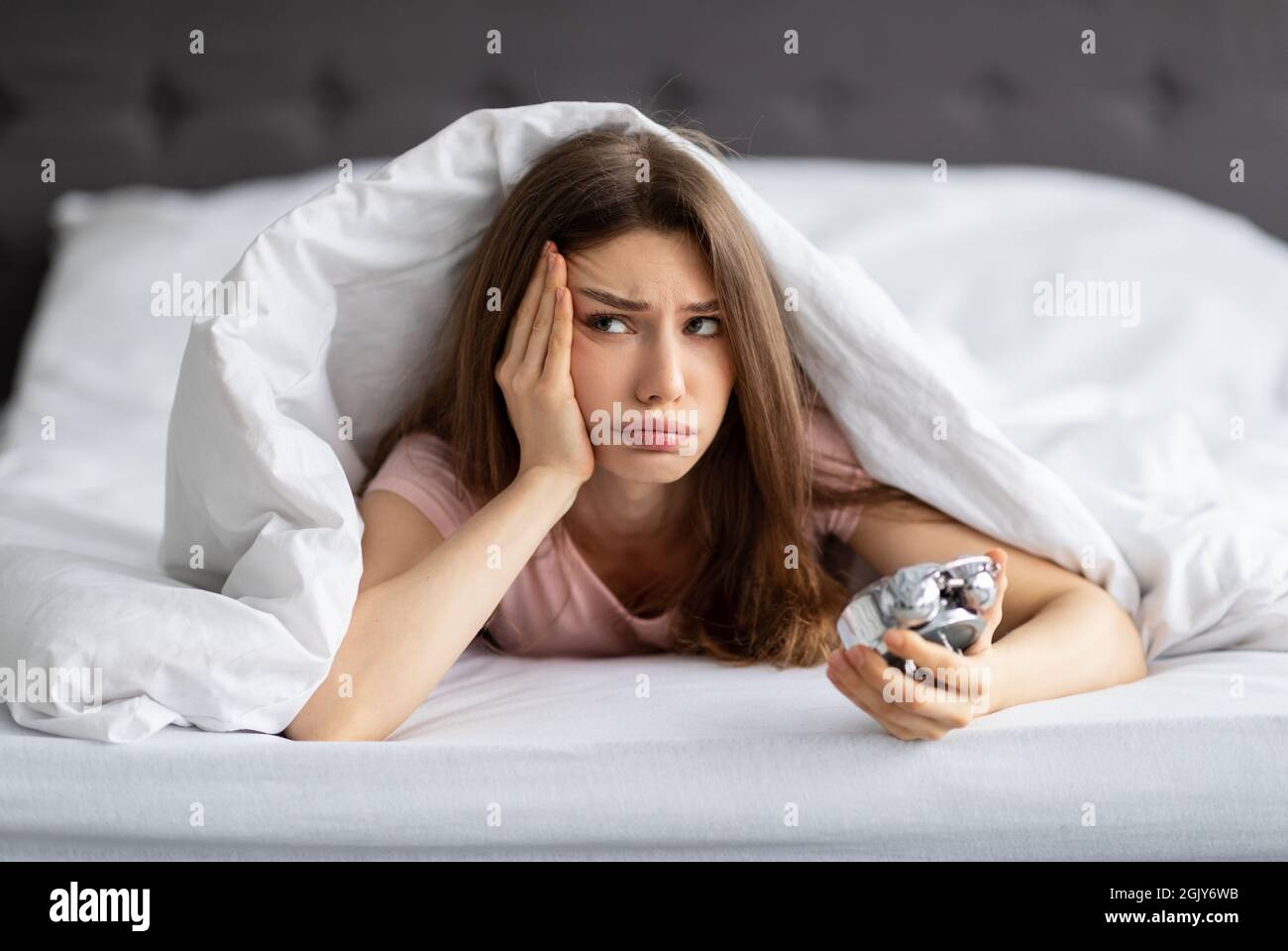 Infelice giovane donna caucasica sdraiata a letto con sveglia, non volendo alzarsi al mattino Foto Stock