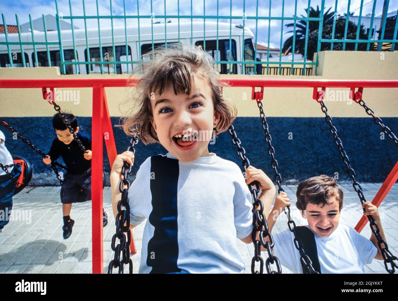 Eccitata bambina in uniforme scolastica 4-6 anni che gioca molto felicemente sulle altalene scolastiche in tempo di pausa, all'aperto al sole con gli altri allievi della scuola Foto Stock