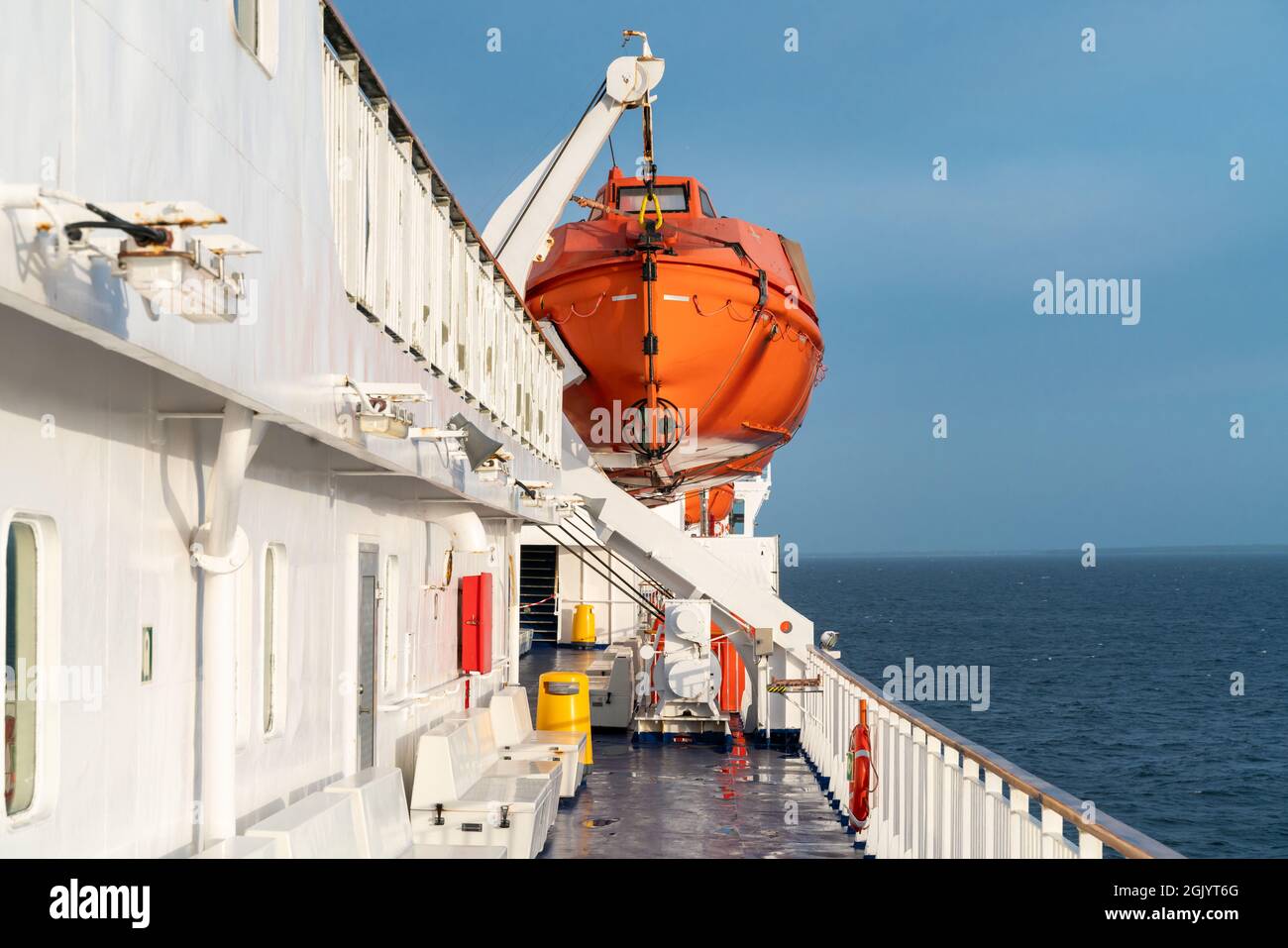 Orange vita barca appesa su una gru su un ponte di barca a vela oceano con orizzonte oceano nella parte posteriore e ringhiera di legno di fronte. Foto Stock
