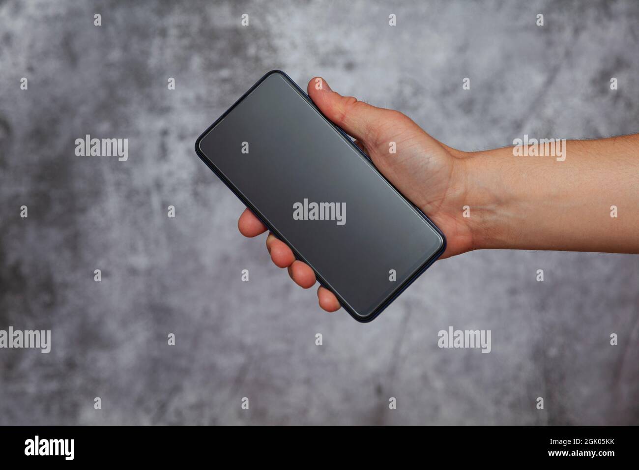 Mano destra di un uomo che tiene uno smartphone con lo schermo spento su uno sfondo grigio testurizzato. Foto Stock