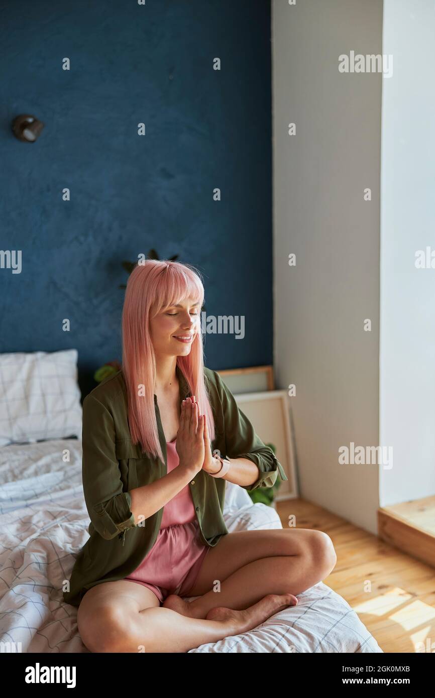 Donna calma con lunghi meditati rosa dei capelli che tengono la mano sin namaste sul letto grande Foto Stock