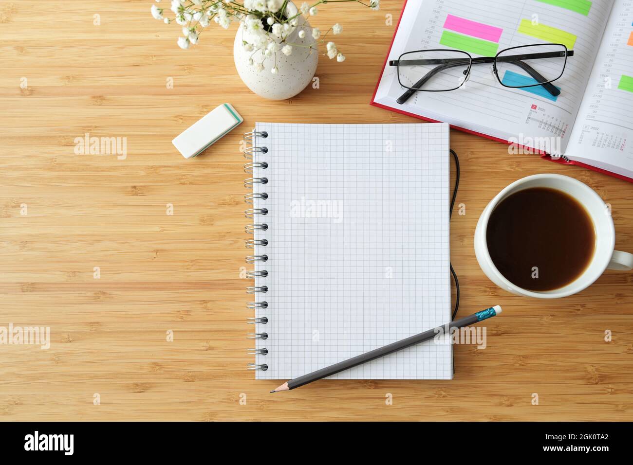 Modello piatto di un notebook a spirale vuoto su una scrivania in legno con organiser, caffè, bicchieri e fiori, spazio copia, focus selezionato, profondità ridotta di Foto Stock