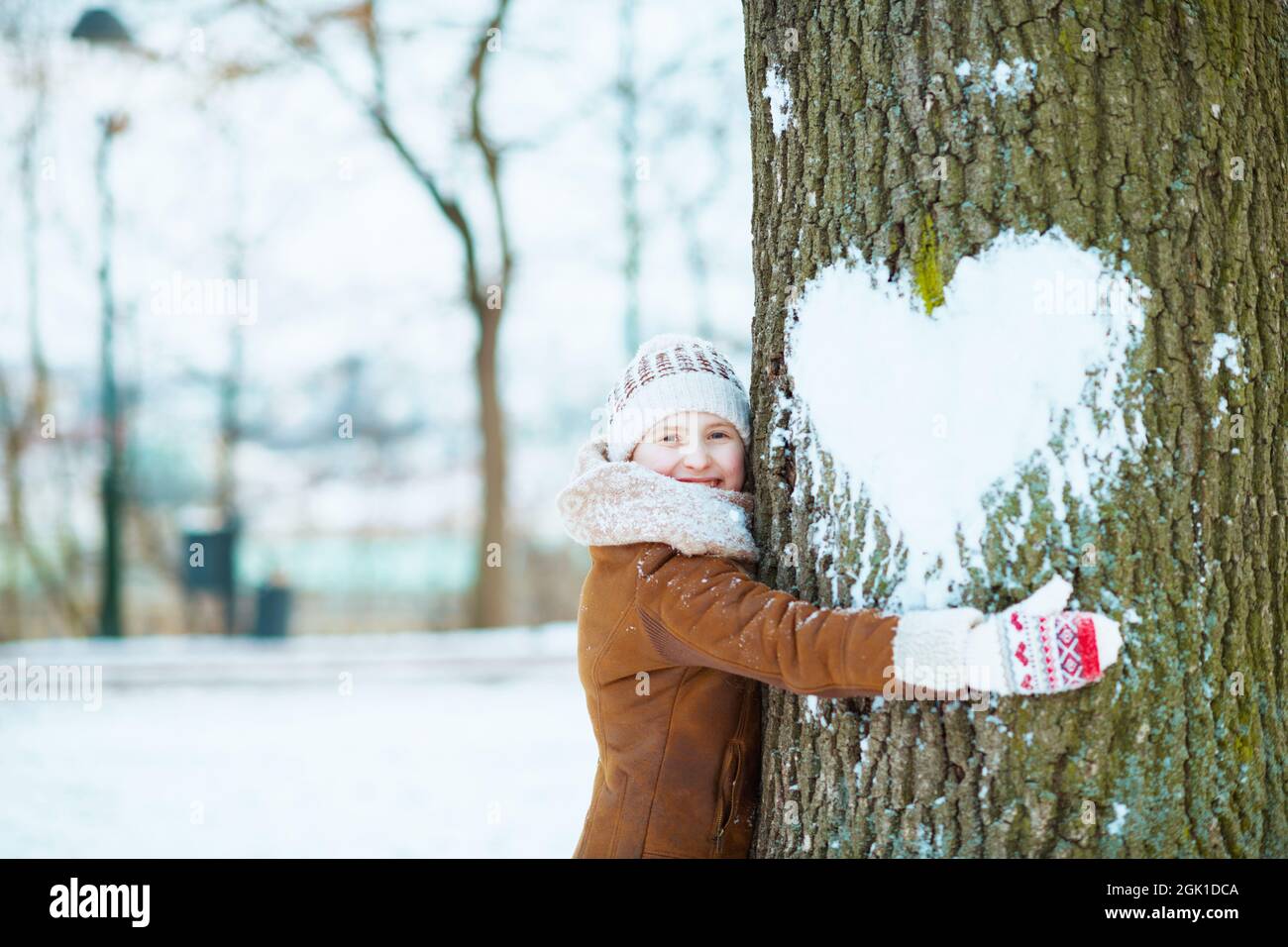 bambino moderno felice con i guanti in un cappello a maglia e cappotto di pelle di pecora albero abbracciare con cuore nevoso fuori nel parco della città in inverno. Foto Stock