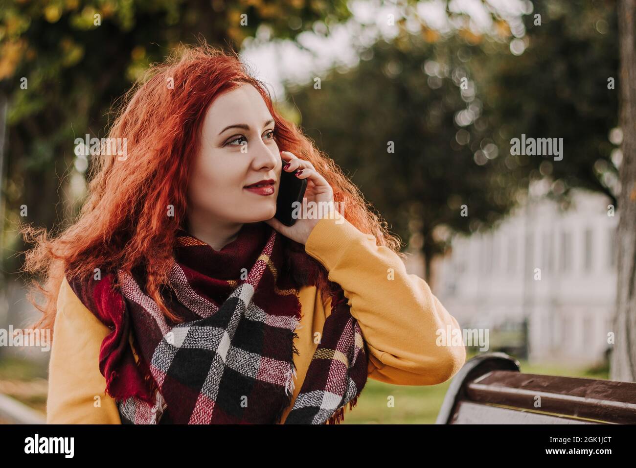 Giovane donna dai capelli rossi in felpa gialla che parla al telefono. Lady siede sul banco del parco il giorno dell'autunno. Stile di vita. Foto Stock