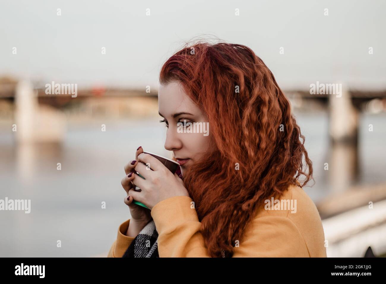 Ritratto di giovane donna dai capelli rossi sullo sfondo del fiume e del ponte della città. Beve caffè da una tazza di carta e gode di una vista autunnale. Spazio di copia. Foto Stock