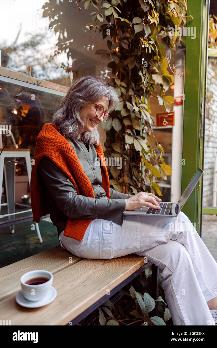 Donna matura felice con i tipi di occhiali su tastiera portatile seduta su una vetrina caffè Foto Stock