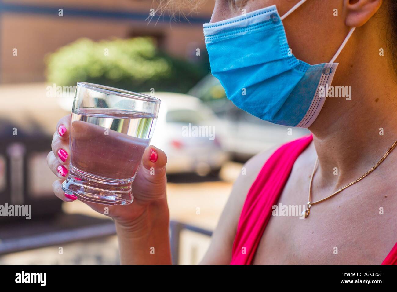 Ragazza in blu maschera medica all'aperto tiene in mano un bicchiere di acqua ancora pura. Bere alcolici nel periodo post pandemico. Indossare una maschera protettiva Foto Stock