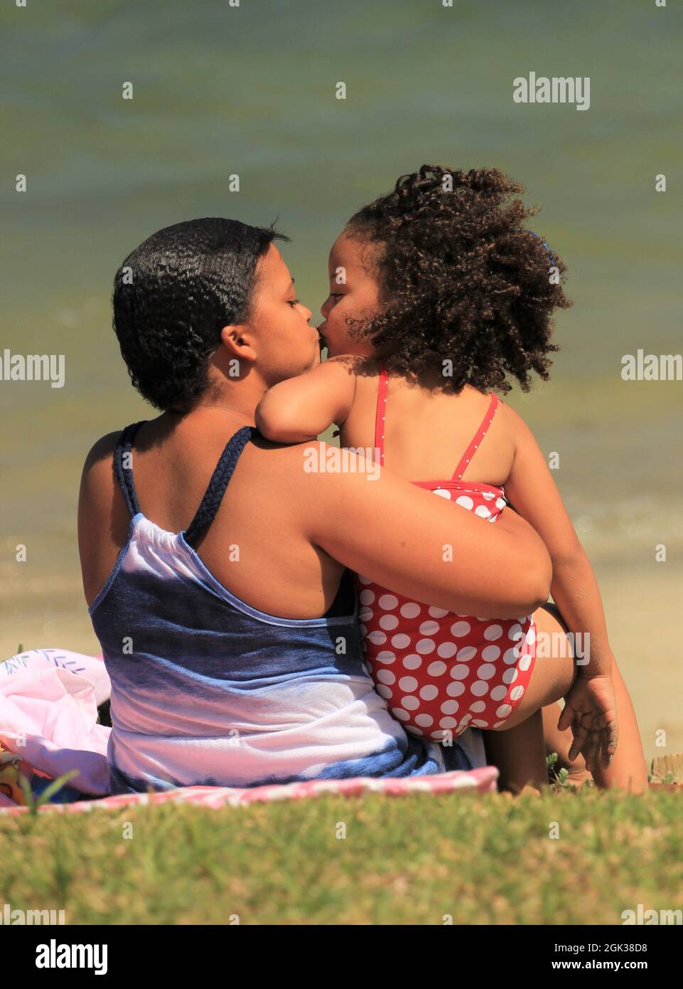 ute piccola ragazza africana con capelli ricci e sua madre seduta su una spiaggia e abbracciarsi l'un l'altro Foto Stock