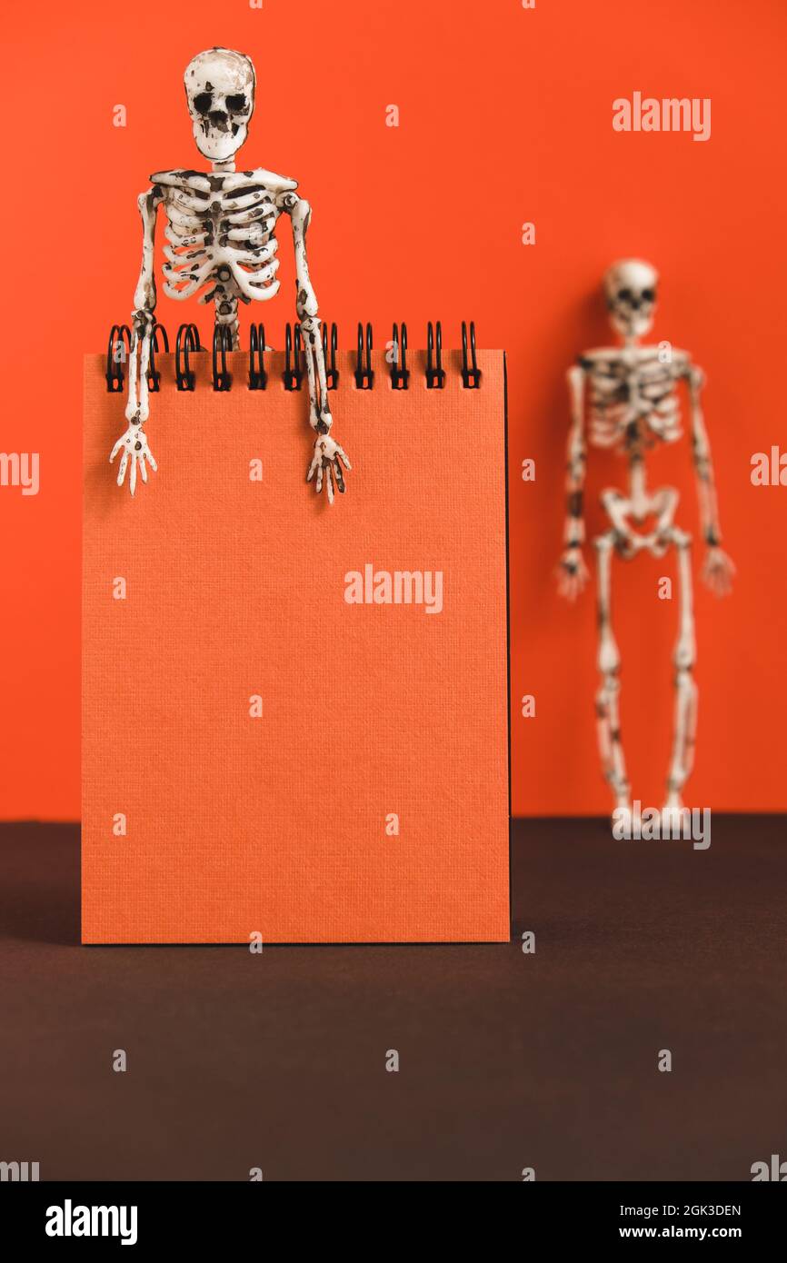 Notebook arancione vuoto su sfondo arancione con scheletri. Design minimalista e luminoso per la preparazione di Halloween. Tessera vacanza. Copia spase. Foto Stock