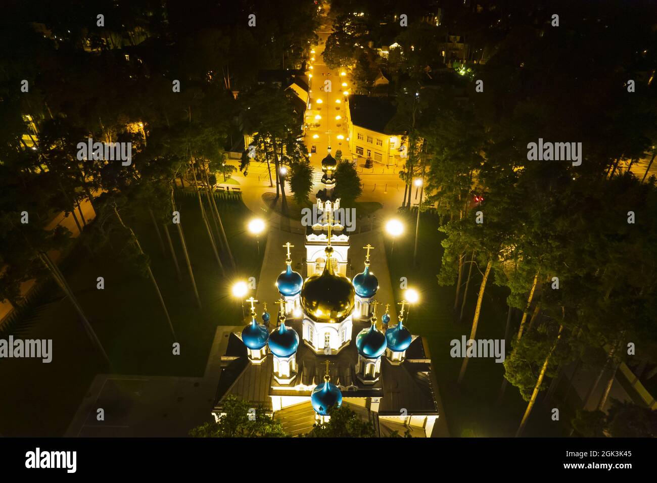 Vista aerea della chiesa illuminata tra i pini di notte. Nuova cappella pubblica vuota bianca con cupole dorate e blu all'aperto dall'alto. Bel luogo religioso, sacro per i ministeri Foto Stock
