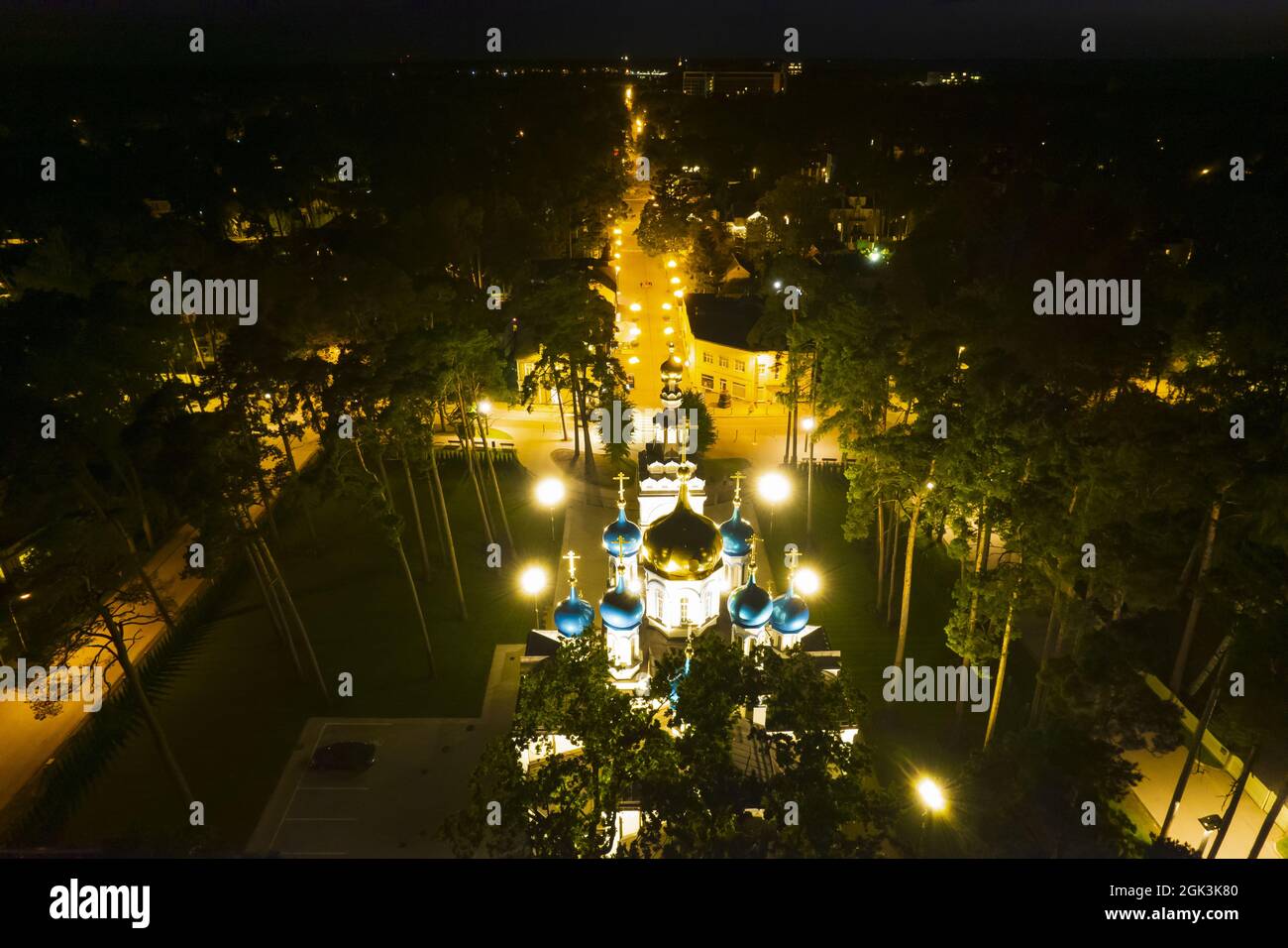 Vista aerea della chiesa illuminata tra i pini di notte. Nuova cappella pubblica vuota bianca con cupole dorate e blu all'aperto dall'alto. Bel luogo religioso, sacro per i ministeri Foto Stock